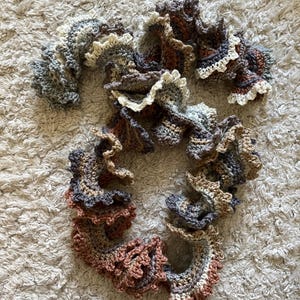 May include: A handmade, ruffled crochet scarf in shades of gray, brown, and cream. The scarf has a wavy, textured design with a layered effect. The yarn appears soft and the scarf is lying on a neutral-colored, textured surface.