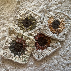 May include: Four crocheted squares with a circular design in the center. The squares are made with a variety of colors, including beige, brown, green, orange, and red. The squares are arranged in a semi-circle on a white fluffy surface.