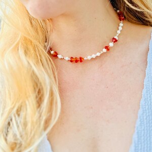 Puede incluir: Un collar delicado con perlas blancas y cuentas de piedras preciosas rojas. El collar está puesto en el cuello de una persona.