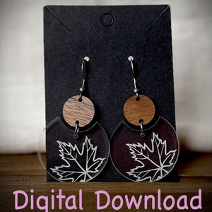 May include: A pair of dangle earrings with a wood circle and a clear acrylic circle with a white maple leaf design. The earrings are hanging on a black display card.