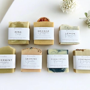 May include: An overhead shot of eight handcrafted natural soaps with white labels. The soaps are labeled with scents like Pine Forest, Orange Grapefruit, and Lemon Poppyseed. Each soap is approximately 7.6 cm wide and 5 cm tall. White flowers and a dried orange slice are also in the image.