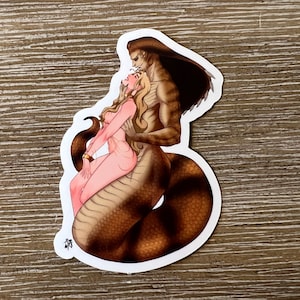 May include: A sticker featuring a fantasy illustration of a humanoid figure with a snake tail embracing a woman. The figures are in shades of brown and pink, with the woman having long blonde hair. The sticker has a white border.