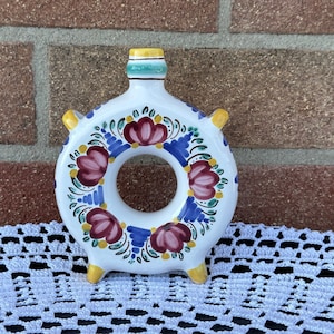 Könnte beinhalten: Eine weiße, ringförmige Flasche aus Keramik mit einem Blumendesign in Rot-, Blau-, Gelb- und Grüntönen. Die Flasche hat eine kleine Öffnung oben und vier kleine Füße.