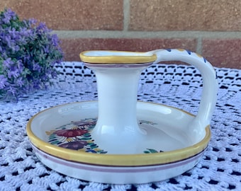 May include: White ceramic candle holder with a yellow and pink floral design and a handle. The holder is on a white crocheted surface.