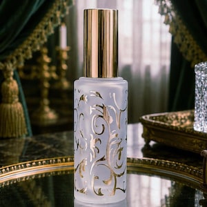 May include: A cylindrical frosted glass spray bottle featuring a metallic gold cap and a decorative gold filigree pattern of swirling vines and leaves printed on the exterior surface of the glass container, standing upright on a reflective surface.