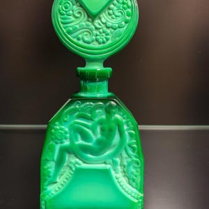 Puede incluir: Un frasco de perfume de vidrio verde vibrante con un tap&oacute;n circular grande que presenta intrincados relieves florales. El cuerpo rectangular muestra un relieve moldeado de una figura sentada rodeada de patrones bot&aacute;nicos y bordes decorativos.