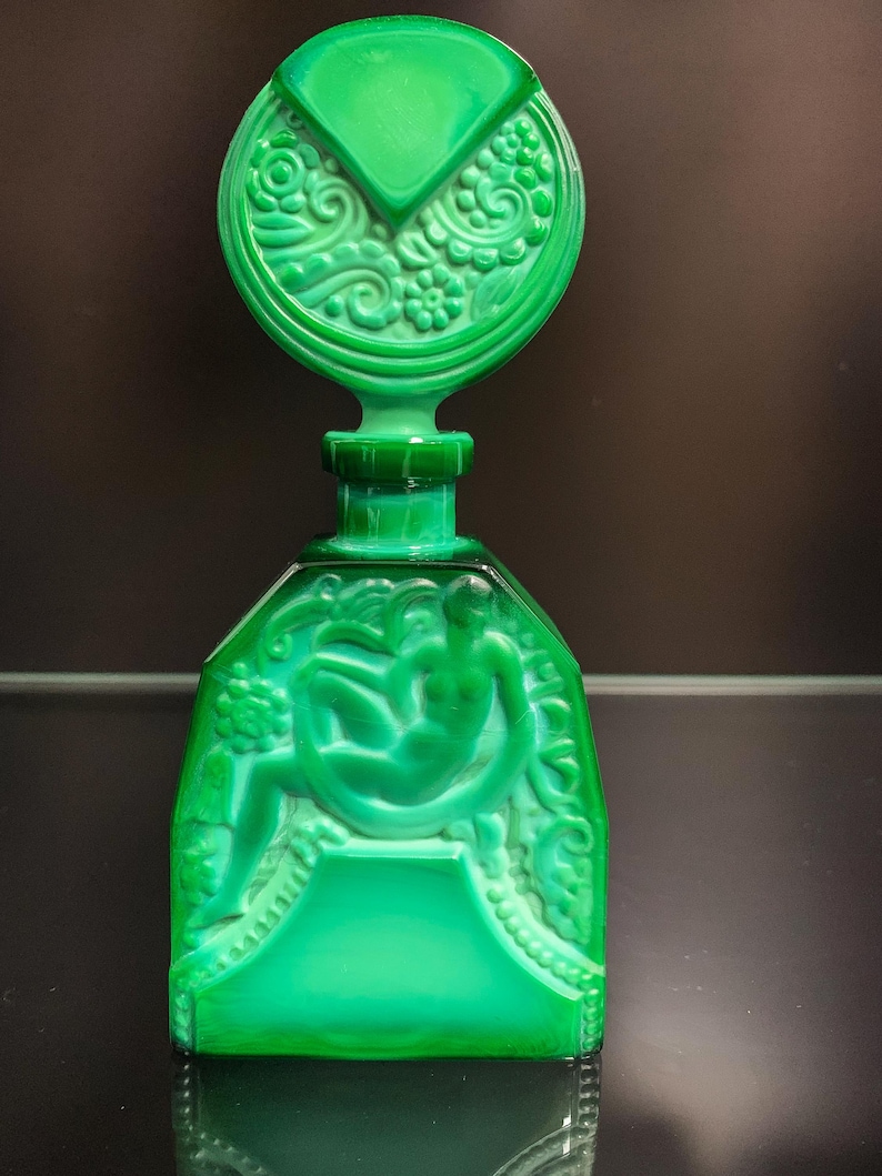 Puede incluir: Un frasco de perfume de vidrio verde vibrante con un tap&oacute;n circular grande que presenta intrincados relieves florales. El cuerpo rectangular muestra un relieve moldeado de una figura sentada rodeada de patrones bot&aacute;nicos y bordes decorativos.