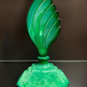 Puede incluir: Un frasco de perfume de vidrio verde vibrante con una base geom&eacute;trica facetada con un patr&oacute;n jaspeado y un tap&oacute;n escultural alto con forma de hoja curva estilizada, con crestas distintas que se extienden desde el centro al borde.