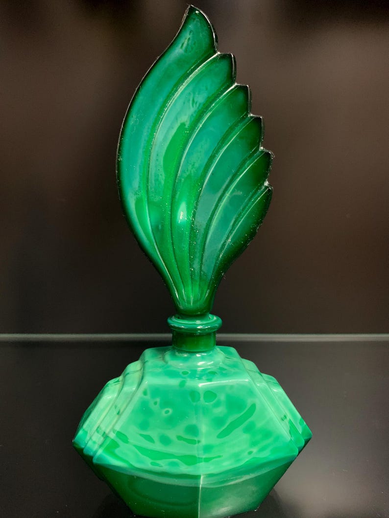 Puede incluir: Un frasco de perfume de vidrio verde vibrante con una base geom&eacute;trica facetada con un patr&oacute;n jaspeado y un tap&oacute;n escultural alto con forma de hoja curva estilizada, con crestas distintas que se extienden desde el centro al borde.