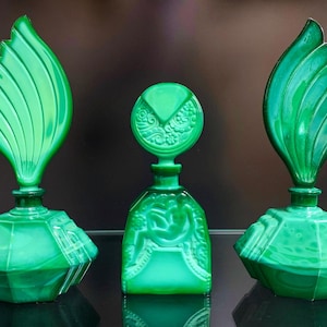 Puede incluir: Tres frascos de perfume de vidrio verde con apariencia de malaquita. Dos frascos presentan tapones grandes en forma de hoja con detalles acanalados, mientras que el frasco central tiene una base cuadrada y un tap&oacute;n redondo con relieves florales.