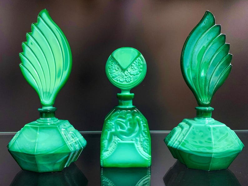 Puede incluir: Tres frascos de perfume de vidrio verde con apariencia de malaquita. Dos frascos presentan tapones grandes en forma de hoja con detalles acanalados, mientras que el frasco central tiene una base cuadrada y un tap&oacute;n redondo con relieves florales.