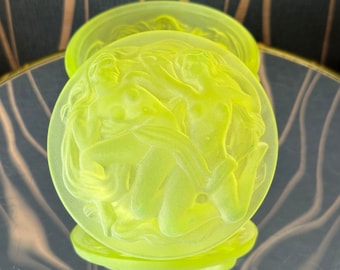 May include: A set of three round, neon-yellow glass coasters. Each coaster features a bas-relief design of two figures in an embrace. The coasters are displayed on a reflective surface, enhancing the glow of the glass.