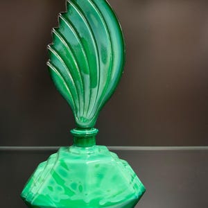 Puede incluir: Un frasco de perfume de vidrio verde con una base hexagonal facetada y un tap&oacute;n alto en forma de hoja con estr&iacute;as verticales. El vidrio presenta un patr&oacute;n veteado semitransl&uacute;cido en varios tonos de verde esmeralda y verde bosque.