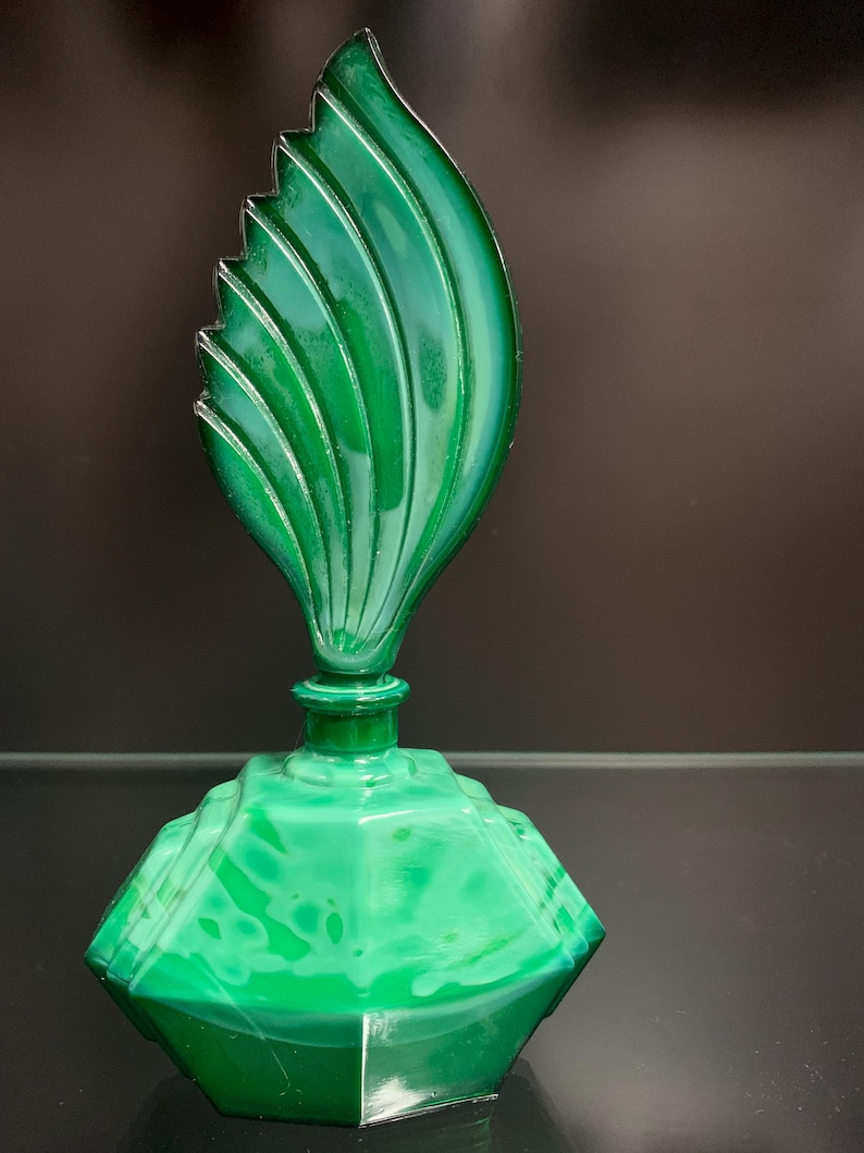 Puede incluir: Un frasco de perfume de vidrio verde con una base hexagonal facetada y un tap&oacute;n alto en forma de hoja con estr&iacute;as verticales. El vidrio presenta un patr&oacute;n veteado semitransl&uacute;cido en varios tonos de verde esmeralda y verde bosque.