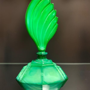Puede incluir: Un frasco de perfume de vidrio verde vibrante con una base hexagonal facetada y un tap&oacute;n escult&oacute;rico alto con forma de llama o hoja estilizada con crestas verticales. El vidrio muestra una apariencia semitransl&uacute;cida y veteada en todo el cuerpo.