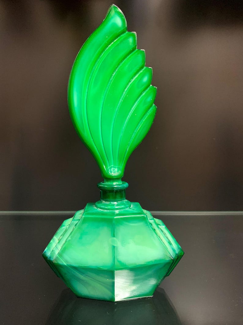 Puede incluir: Un frasco de perfume de vidrio verde vibrante con una base hexagonal facetada y un tap&oacute;n escult&oacute;rico alto con forma de llama o hoja estilizada con crestas verticales. El vidrio muestra una apariencia semitransl&uacute;cida y veteada en todo el cuerpo.