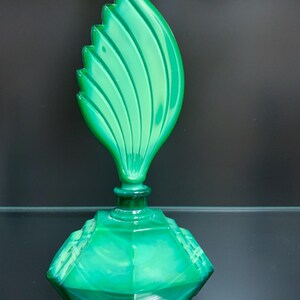 Puede incluir: Un frasco de perfume de vidrio verde vibrante con una base facetada de varios lados y un tap&oacute;n escultural alto con forma de hoja estilizada con crestas verticales. El vidrio muestra una apariencia transl&uacute;cida y veteada con tonos verdes.