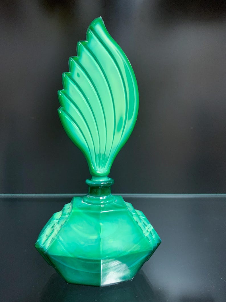 Puede incluir: Un frasco de perfume de vidrio verde vibrante con una base facetada de varios lados y un tap&oacute;n escultural alto con forma de hoja estilizada con crestas verticales. El vidrio muestra una apariencia transl&uacute;cida y veteada con tonos verdes.