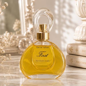 Puede incluir: Un frasco de perfume de vidrio transparente lleno de líquido dorado, con un cuello de tono dorado y un tapón redondeado y transparente. La etiqueta muestra "Eau de Toilette", "First" y "Van Cleef & Arpels" en texto negro en el frente.