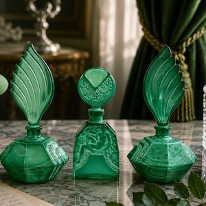 Puede incluir: Tres frascos de perfume de vidrio verde esmeralda. Dos frascos tienen bases anchas facetadas y tapones altos en forma de hoja. El frasco central es rectangular con una figura en relieve y un tap&oacute;n circular con patrones florales.