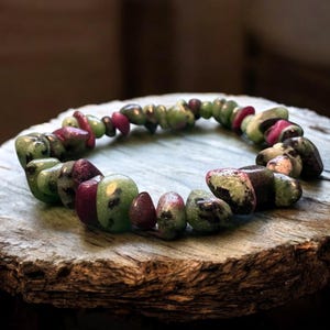 May include: A bracelet made from a series of polished, irregularly shaped stones. The stones are predominantly green and burgundy, with some showing a mottled pattern. The bracelet is presented on a rustic wooden surface, highlighting its natural appeal.