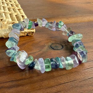 May include: A bracelet composed of polished fluorite stones in shades of green, purple, and clear. The stones are strung together, forming a circular shape. The bracelet is displayed on a wooden surface.