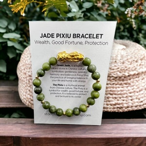 May include: A green jade Pixiu bracelet with gold detailing. The bracelet has round green jade beads and a gold Pixiu charm. The card behind the bracelet reads "JADE PIXIU BRACELET Wealth, Good Fortune, Protection."