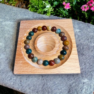K&ouml;nnte beinhalten: Ein Armband aus bunten Edelsteinperlen in Braun-, Gr&uuml;n- und Graut&ouml;nen. Das Armband liegt auf einem quadratischen Holztablett mit einer kreisf&ouml;rmigen Vertiefung. Das Tablett ist aus hellem Holz gefertigt.