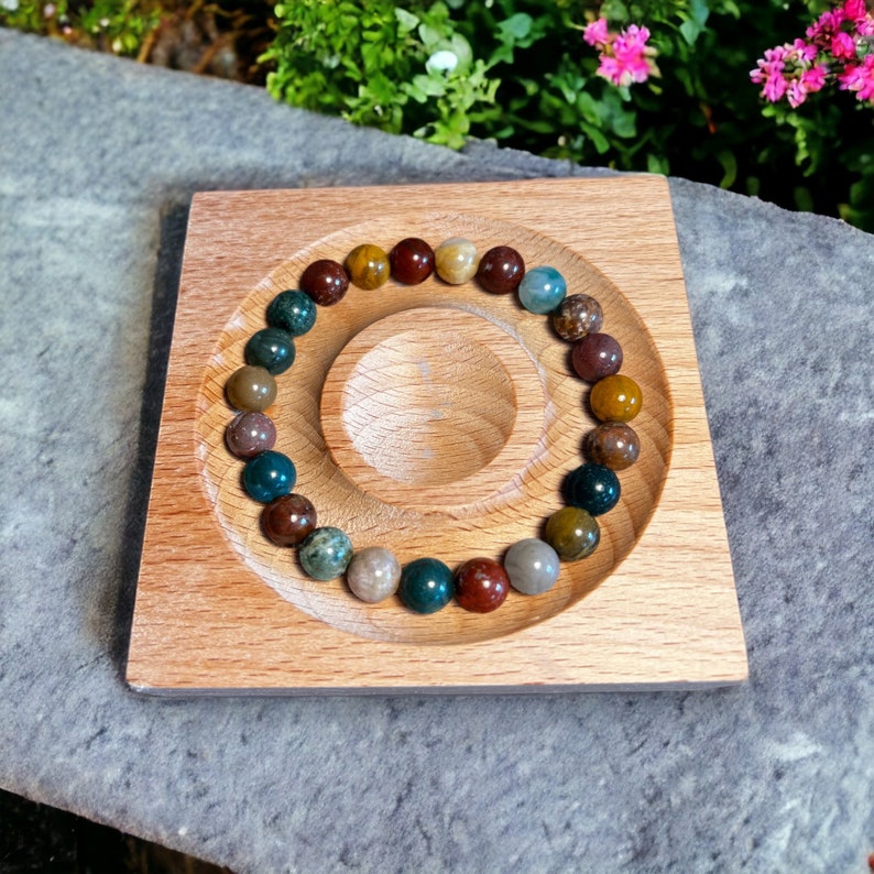 K&ouml;nnte beinhalten: Ein Armband aus bunten Edelsteinperlen in Braun-, Gr&uuml;n- und Graut&ouml;nen. Das Armband liegt auf einem quadratischen Holztablett mit einer kreisf&ouml;rmigen Vertiefung. Das Tablett ist aus hellem Holz gefertigt.