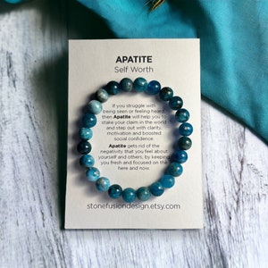 May include: Apatite gemstone bracelet with a card that reads "APATITE Self Worth". The bracelet features round, blue-green beads. The card includes text about the benefits of Apatite for self-worth and clarity. The background is a light wood surface.