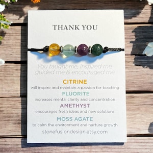 May include: A bracelet featuring four round gemstone beads in yellow, light green, purple, and dark green, separated by small silver-toned metal spacers on a black cord. The bracelet is attached to a white card with the text THANK YOU and descriptions of citrine, fluorite, amethyst, and moss agate.