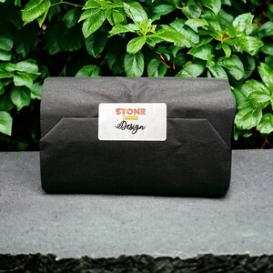 K&ouml;nnte beinhalten: Ein in schwarzes Papier verpacktes Paket mit einem wei&szlig;en Etikett mit der Aufschrift "STONE FUSION Design". Das Paket liegt auf einer grauen Oberfl&auml;che, mit gr&uuml;nem Laub im Hintergrund.