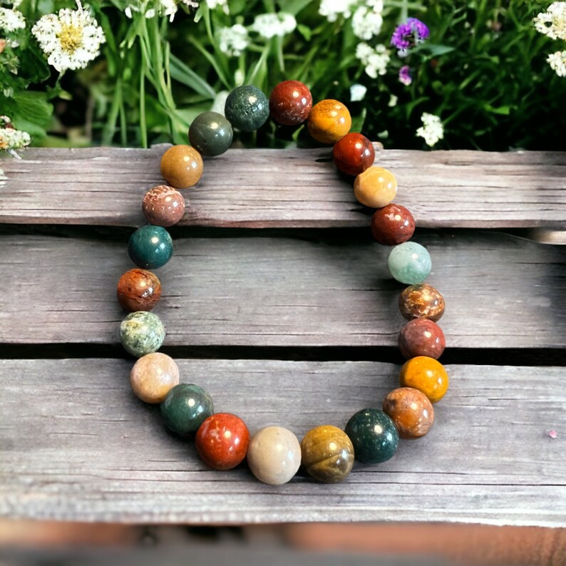 K&ouml;nnte beinhalten: Ein Armband mit bunten, runden Edelsteinen. Die Perlen sind in Rot-, Gr&uuml;n-, Gelb-, Braun- und Cremet&ouml;nen gehalten. Das Armband liegt auf einer verwitterten Holzoberfl&auml;che, mit Gr&uuml;nzeug im Hintergrund.