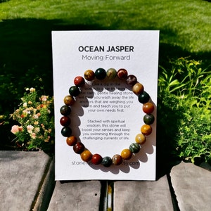 K&ouml;nnte beinhalten: Ein Armband aus Ocean Jasper-Perlen in Gr&uuml;n-, Braun- und Rott&ouml;nen. Das Armband wird mit einer Karte pr&auml;sentiert, die "OCEAN JASPER Moving Forward" tr&auml;gt und die Eigenschaften des Steins beschreibt.