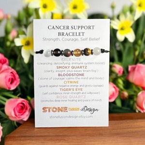May include: A black cord bracelet featuring a selection of gemstone beads: Selenite, Smoky Quartz, Bloodstone, Citrine, Tiger's Eye, and Rose Quartz. The accompanying card states "Cancer Support Bracelet" and stone properties.
