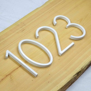 May include: White painted metal house numbers 1023 on a light brown wooden surface.