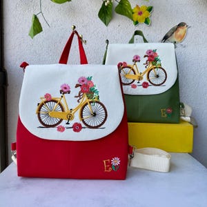 May include: Two embroidered backpacks. One is red with a white flap featuring a yellow bicycle carrying flowers. The other is olive green with a similar design. Both have the letter "E" embroidered on them, and adjustable straps.