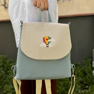 May include: A light blue backpack with a beige flap featuring an embroidered rainbow hot air balloon with a heart. The backpack has adjustable straps.