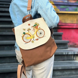 May include: A brown canvas backpack with a beige flap featuring an embroidered image of a yellow bicycle with a basket of flowers. The bicycle is surrounded by yellow leaves.
