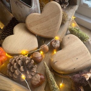 Könnte beinhalten: Drei herzförmige Holzdekorationen in verschiedenen Brauntönen sind mit Tannenzapfen, Kastanien und Lichterketten arrangiert. Die Herzen haben eine glatte Oberfläche und sind auf einer rustikalen Holzoberfläche platziert.