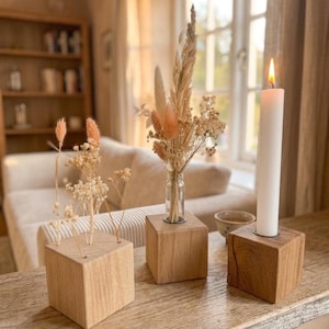 Könnte beinhalten: Drei Kerzenhalter und Vasen aus Holzblöcken auf einer Holzoberfläche. Einer hält eine brennende weiße Kerze, die anderen getrocknete Blumen und Gräser. Die Blöcke sind quadratisch und aus hellem Holz.