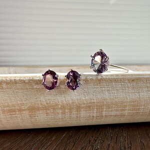 May include: A set of three silver rings with oval-shaped purple gemstones. The rings are on a wooden surface.