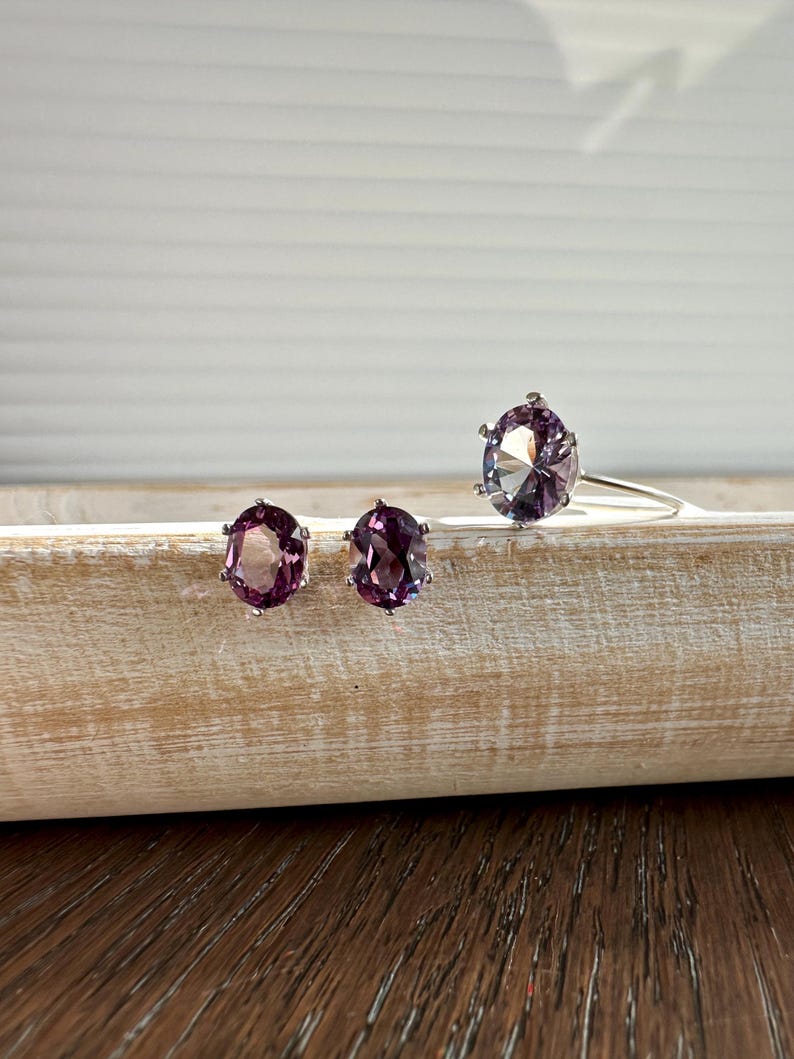 May include: A set of three silver rings with oval-shaped purple gemstones. The rings are on a wooden surface.