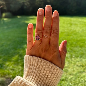 May include: A hand with a light pink nail polish with glitter on the ring finger, wearing a ring with a purple gemstone on the ring finger and two thin gold bands on the middle finger. The hand is wearing a cream colored knitted sweater.