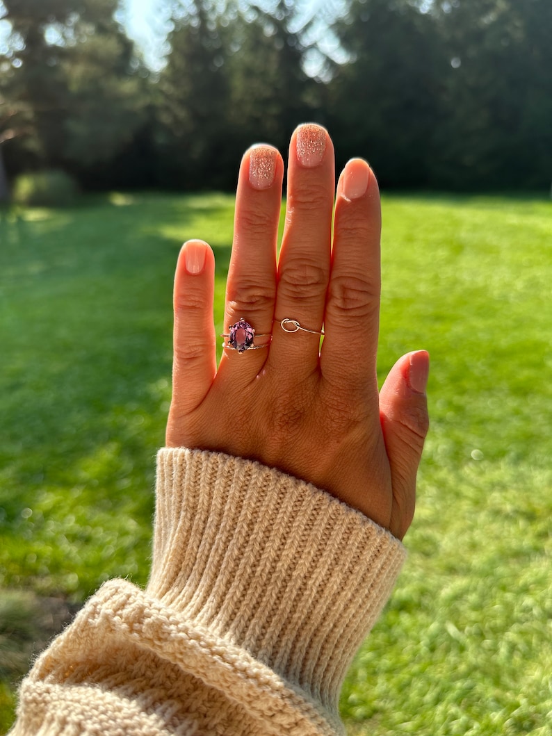 May include: A hand with a light pink nail polish with glitter on the ring finger, wearing a ring with a purple gemstone on the ring finger and two thin gold bands on the middle finger. The hand is wearing a cream colored knitted sweater.