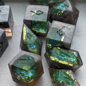 May include: A set of iridescent green and gold dice with black and white markings. The dice are made of resin and have a speckled appearance.