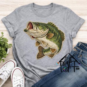 May include: A gray t-shirt featuring a detailed embroidered design of a largemouth bass with an open mouth. The fish is green, brown, and beige. The text "THE CRAFT LOFT" is visible on the lower right.