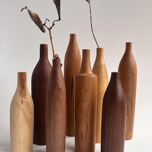 May include: A collection of handcrafted wooden vases in various shades of brown and tan. The vases have a bottle-like shape and are arranged in a group. Some vases have a smooth finish, while others have visible wood grain. Dried plants are placed in some of the vases.