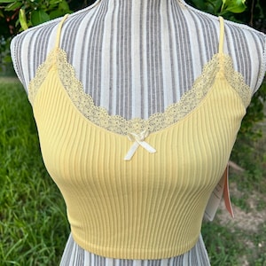 May include: A yellow ribbed crop top with spaghetti straps and a delicate lace trim along the neckline. A small white bow is centered on the front. The top is displayed on a mannequin against a striped background.