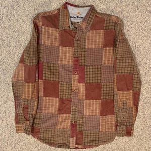 Puede incluir: Una camisa de retazos de manga larga en tonos marrón, rojo y beige. La camisa presenta una parte delantera abotonada, un cuello y una mezcla de cuadrados de tela a cuadros y lisos. La etiqueta dice "Better Vintage".